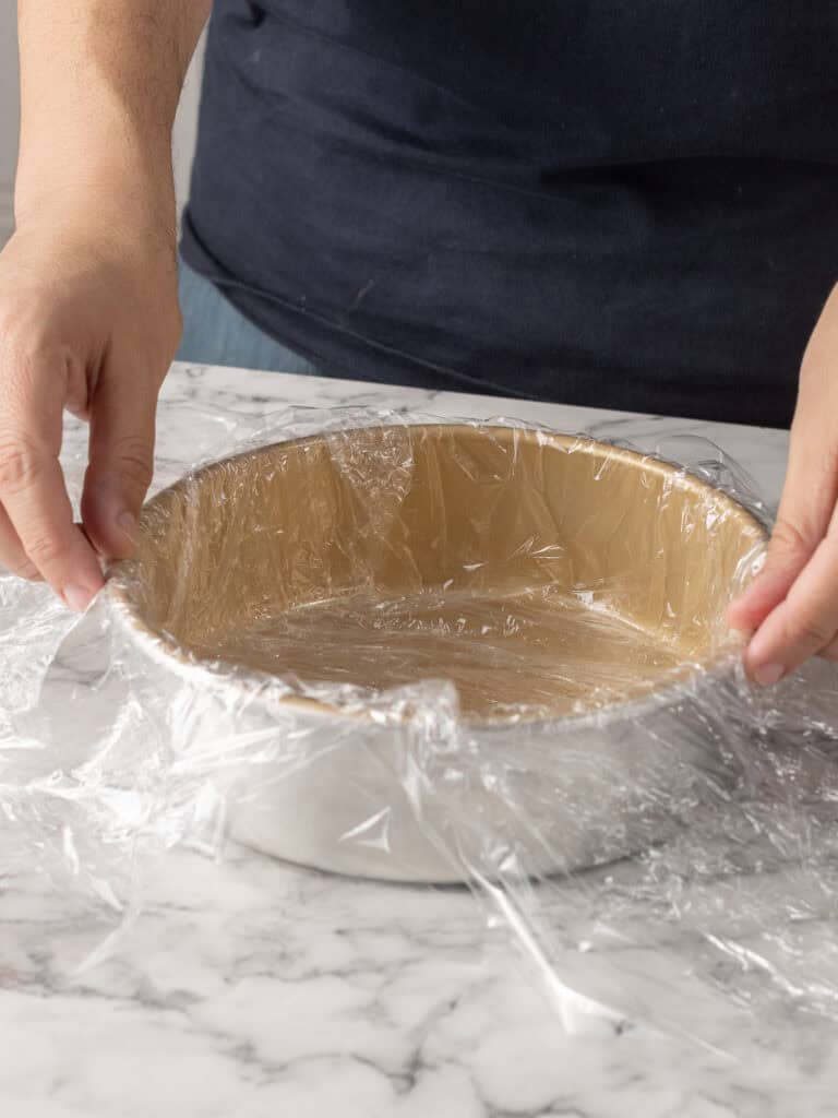 A person is lining a round baking pan with plastic wrap, pressing it against the sides and edges on a marble countertop.