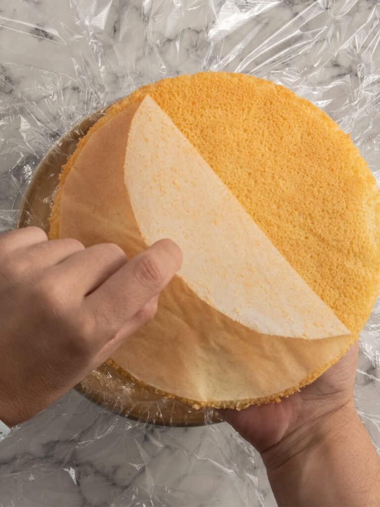 A person peels parchment paper from a round yellow sponge cake placed on plastic wrap over a marble surface.
