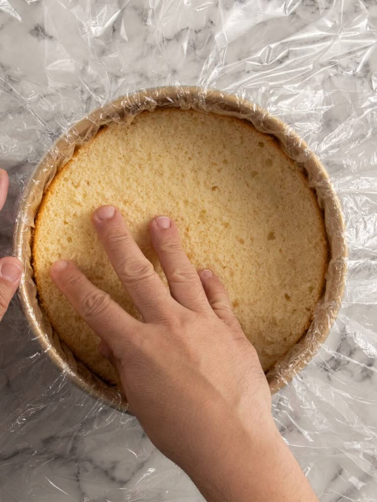 A hand gently presses a round, golden cake layer into a plastic wrap-lined cake pan on a marble surface.