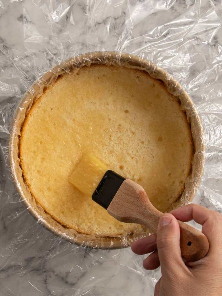 A hand uses a pastry brush to apply syrup to a round sponge cake layer in a pan lined with plastic wrap, on a marble countertop.
