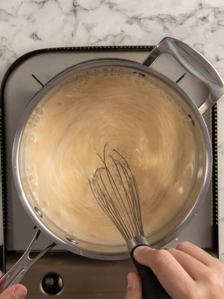 A hand whisking egg yolks and milk to make custard in a saucepan on a stovetop with a marble countertop in the background.