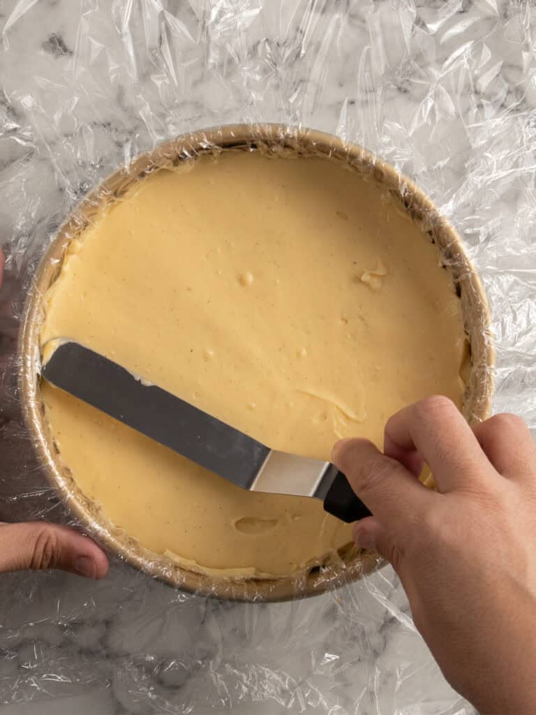A person smooths the surface of a round cheesecake batter in a pan lined with plastic wrap using an offset spatula on a marble countertop.