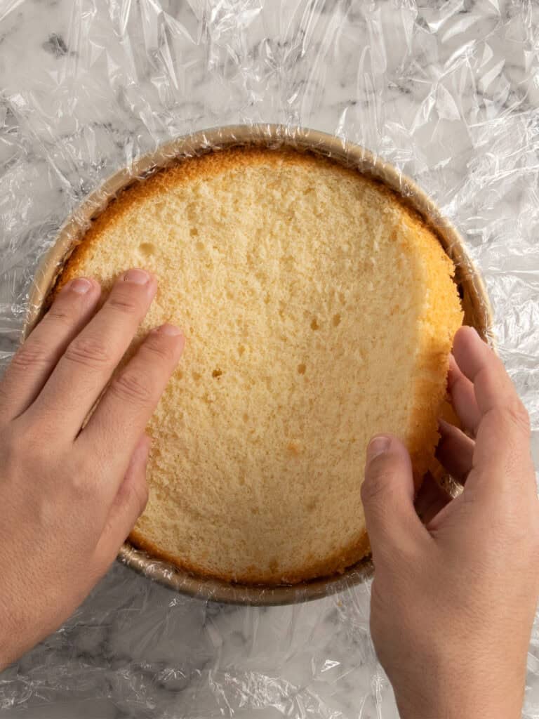 Two hands lift a round, golden sponge cake layer from a cake pan lined with plastic wrap, on a marble surface.