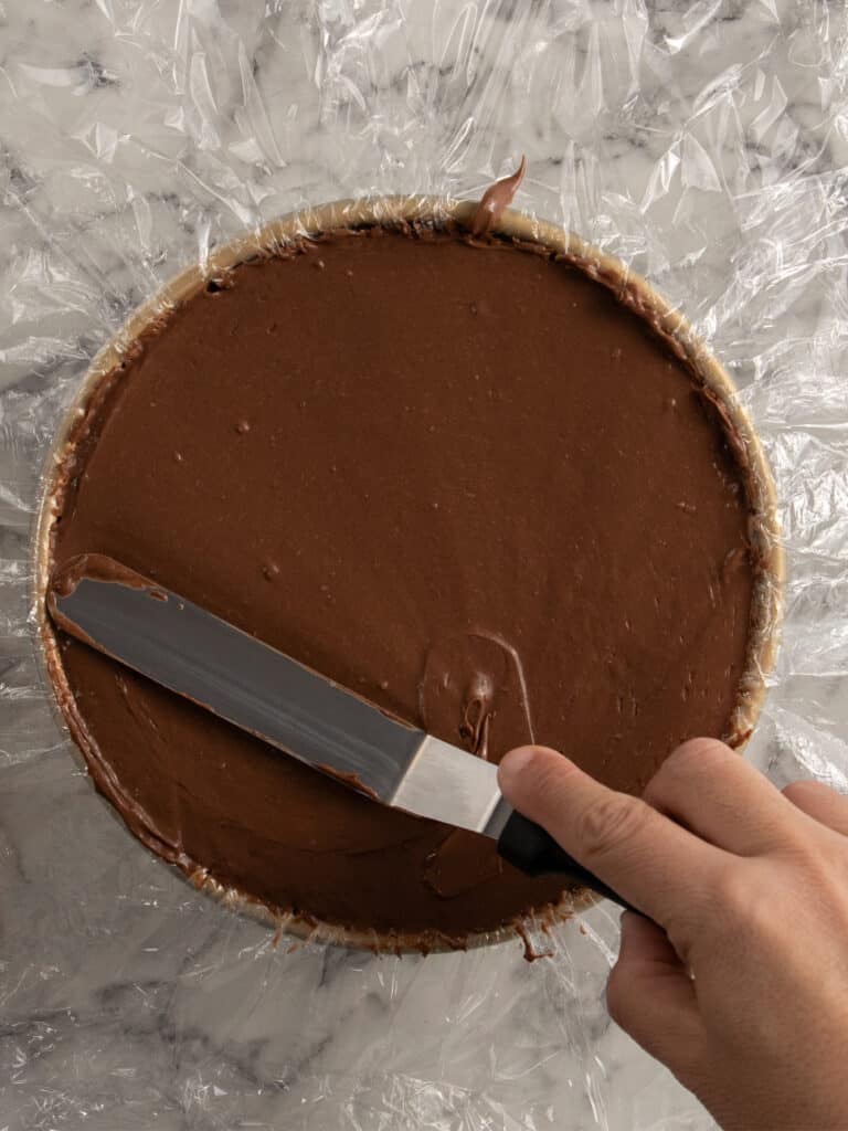 A hand uses an offset spatula to smooth chocolate filling on top of a round dessert, likely a cheesecake or tart, on a sheet of plastic wrap over a marble surface.