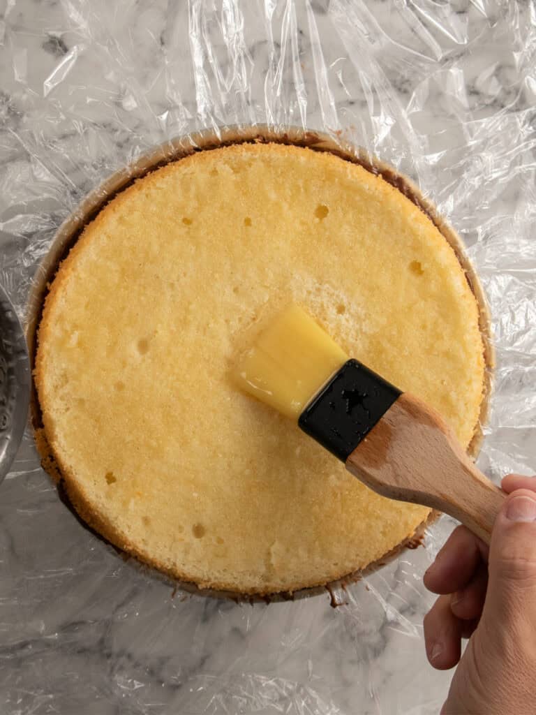 A hand uses a pastry brush to spread syrup on a round, yellow cake layer placed on plastic wrap, with a marble countertop underneath.