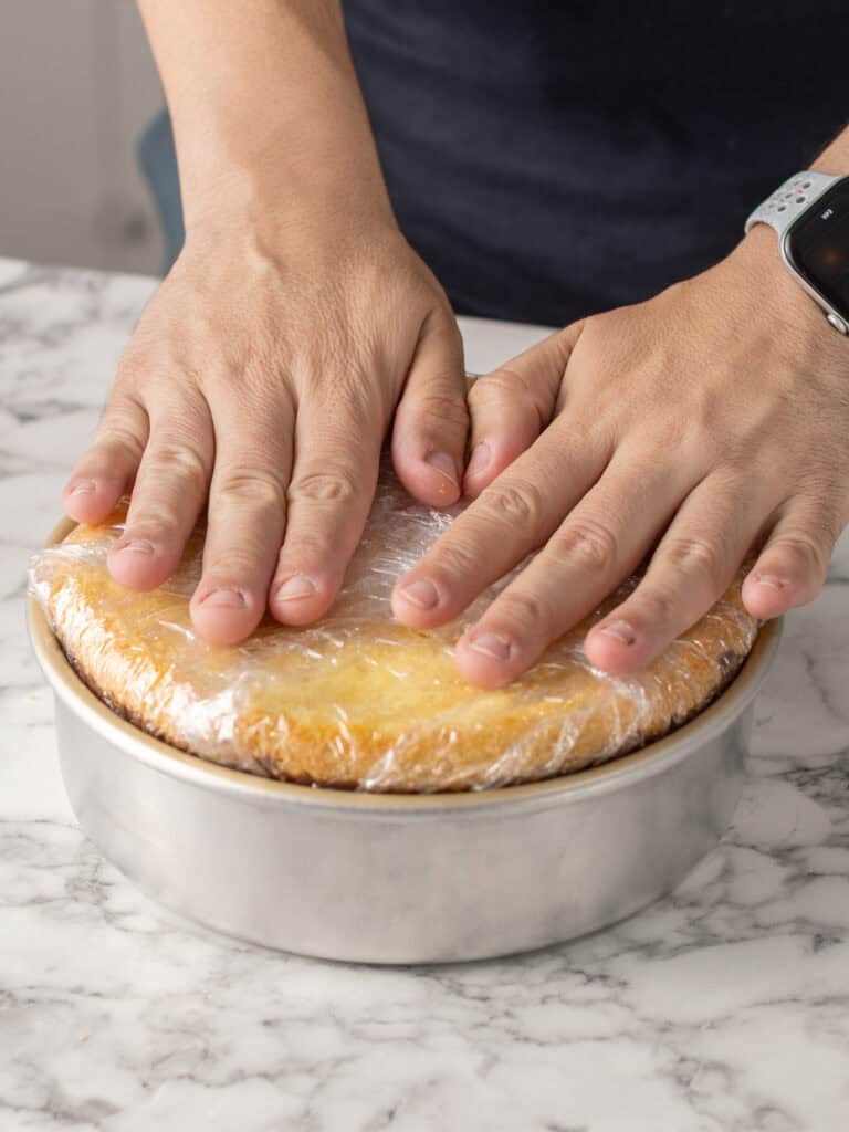 Two hands press plastic wrap over a round yellow cake in a metal pan on a marble countertop. The person is wearing a smartwatch on their left wrist.