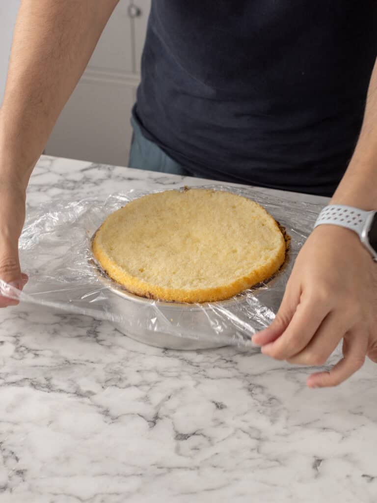 A person covers a round, plain sponge cake on a plate with plastic wrap on a marble countertop.