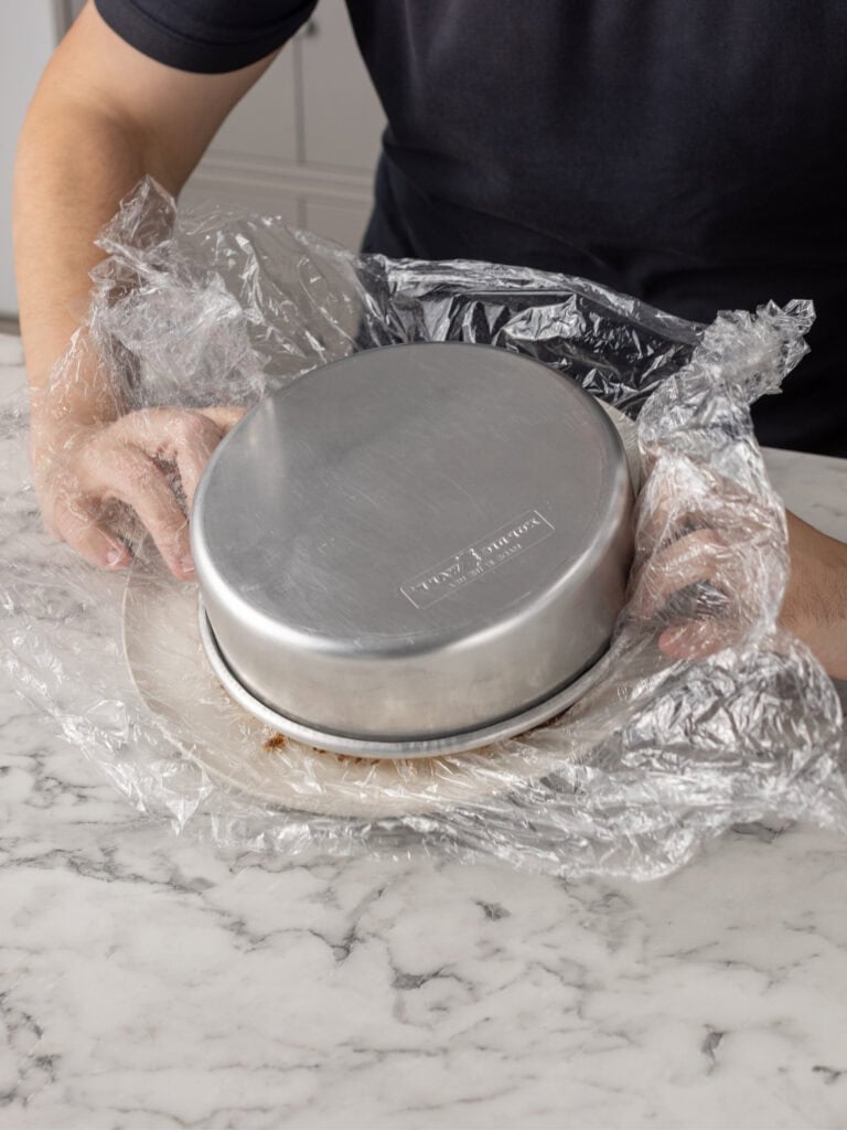 A person wearing a dark shirt removes a metal baking pan from a plate covered in plastic wrap on a marble countertop.