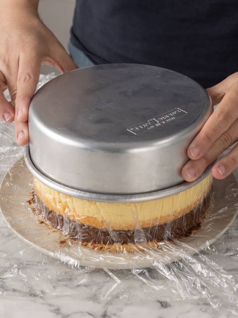 A person lifts a round metal cake pan off a cheesecake covered in plastic wrap, which sits on a marble countertop.