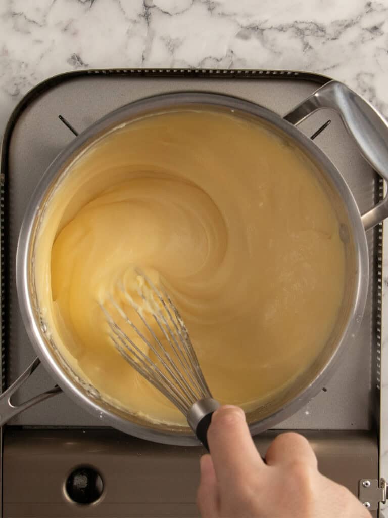 A hand is whisking a thick, pale yellow mixture in a metal saucepan on a stovetop with a marble countertop in the background.