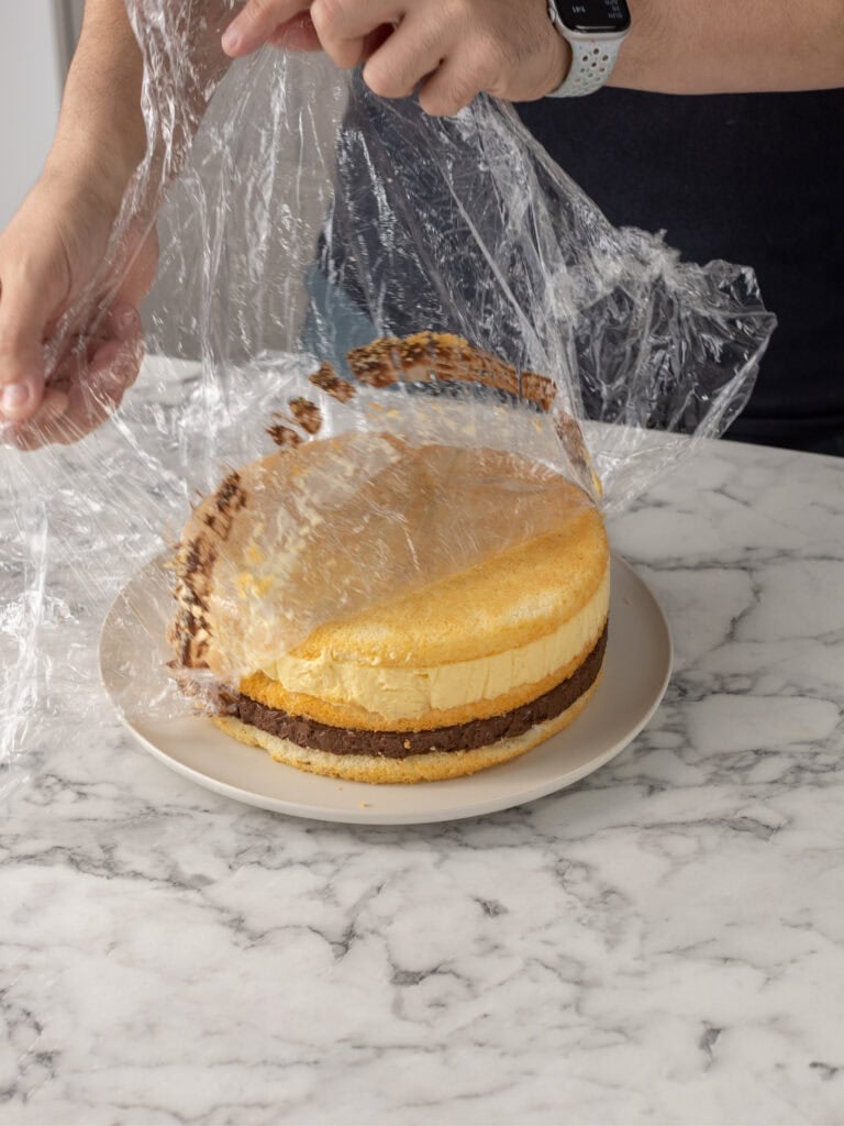 A person’s hands are removing plastic wrap from a round, layered cake on a white plate, set on a marble countertop.