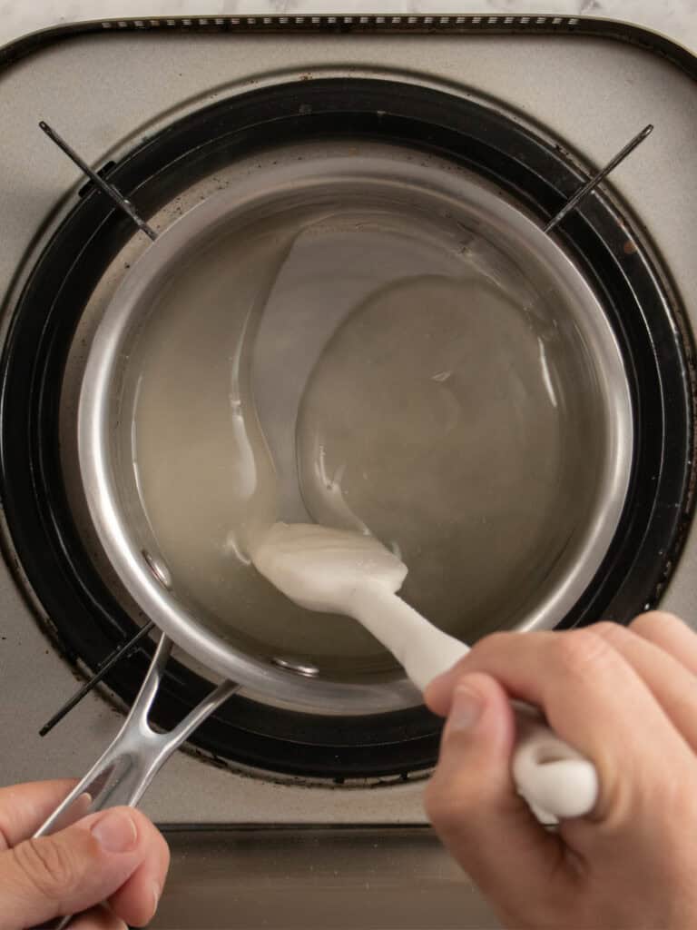 A person stirs a clear, thick liquid in a saucepan on a stove using a white spatula.