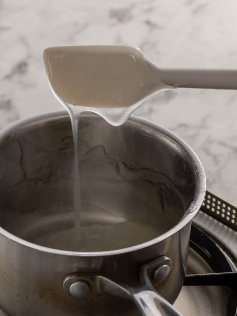 A spatula lifts a thick, clear liquid from a saucepan on a stovetop, with steam rising from the pot. The background features a white, marbled surface.
