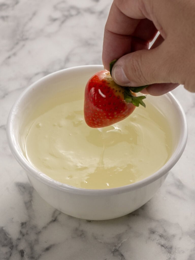 A hand dips a fresh strawberry into a bowl of creamy white chocolate on a marble surface.