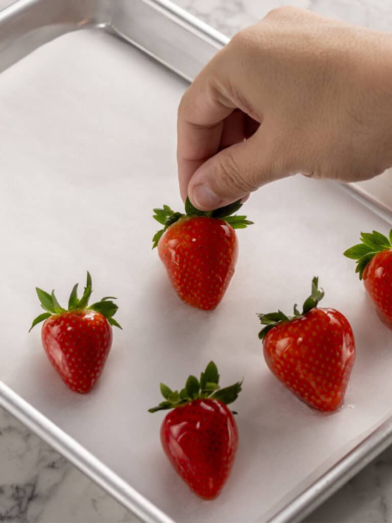 A hand places a fresh strawberry on a parchment-lined baking sheet with four other strawberries, on a light marble surface.