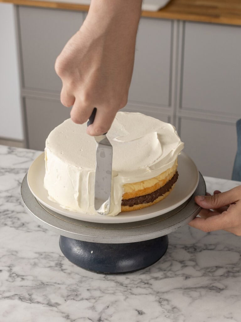 A person uses an offset spatula to spread white frosting on the sides of a layered cake placed on a rotating cake stand on a marble countertop.