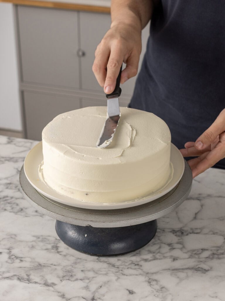A person smooths white frosting on a round cake with an offset spatula, while the cake sits on a stand on a marble countertop.