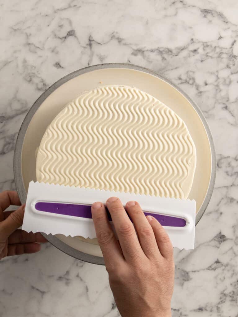 A persons hands use a cake comb to create a wavy textured pattern on the frosting of a round cake sitting on a turntable, with a marble surface in the background.