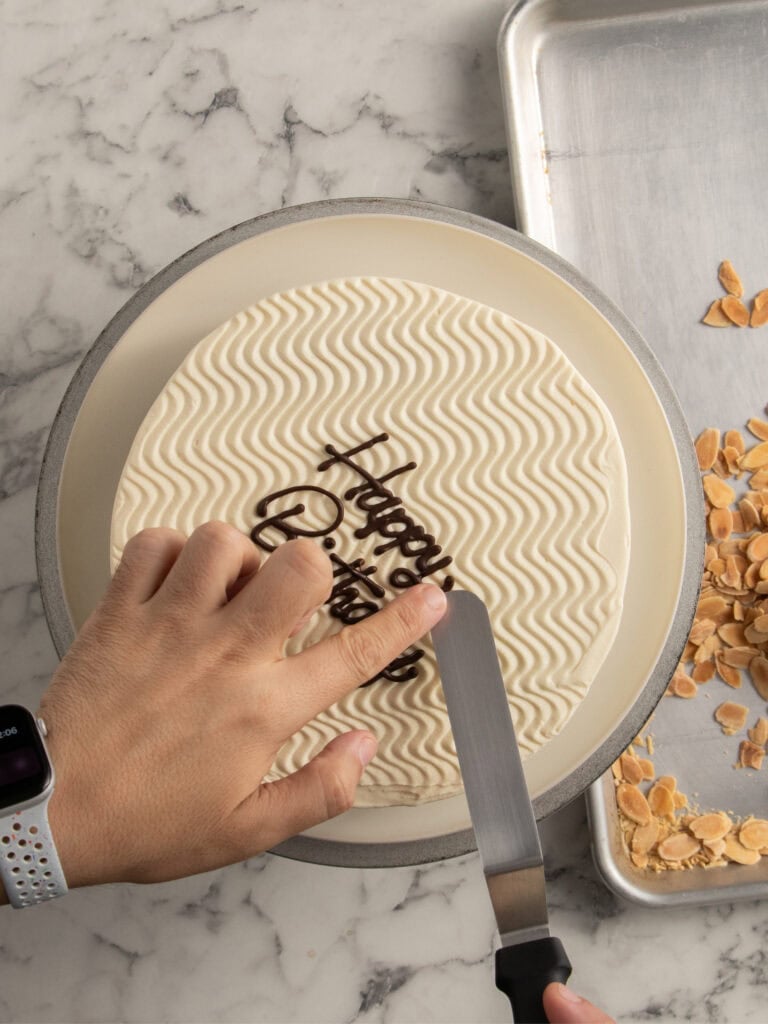 A hand holding a cake with wavy-patterned icing while another hand uses an offset spatula to decorate it with Happy Birthday written in chocolate. Sliced almonds are scattered on a nearby tray.