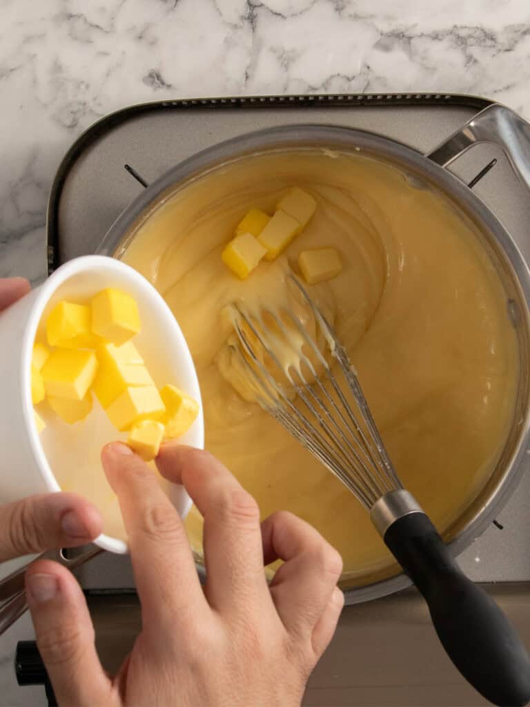 A hand adds cubes of butter from a small bowl into a saucepan of creamy yellow custard being whisked on a stovetop, with a marble countertop in the background.