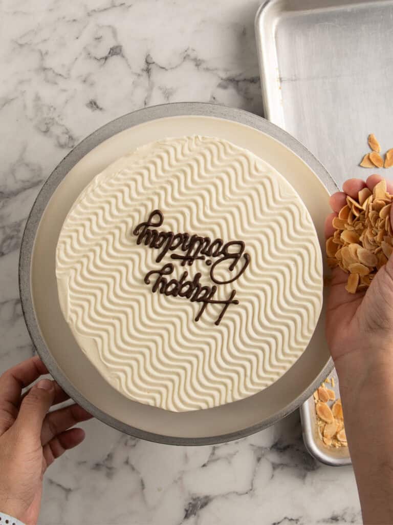A person decorates a round cake with white wavy frosting and chocolate Happy Birthday lettering, holding a handful of sliced almonds next to a metal tray on a marble surface.