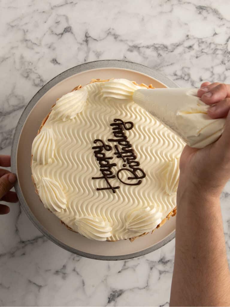 A person decorates a round cake with white frosting and the words Happy Birthday piped in chocolate on top, using a piping bag on a marble countertop.