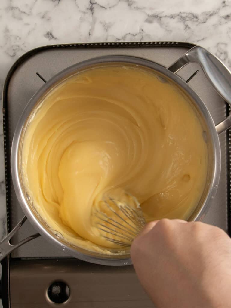 A hand uses a whisk to stir thick, creamy custard in a stainless steel saucepan on a stovetop with a marble countertop background.