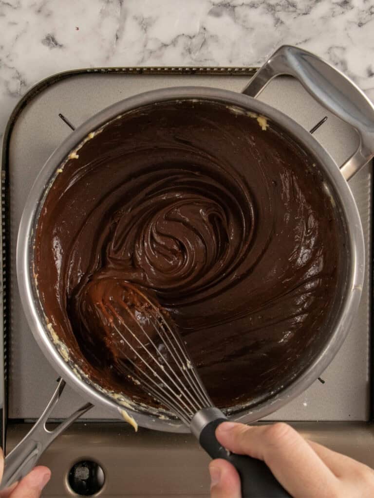 A person whisking smooth, glossy chocolate custard in a saucepan on a stovetop, viewed from above, with a marble countertop in the background.