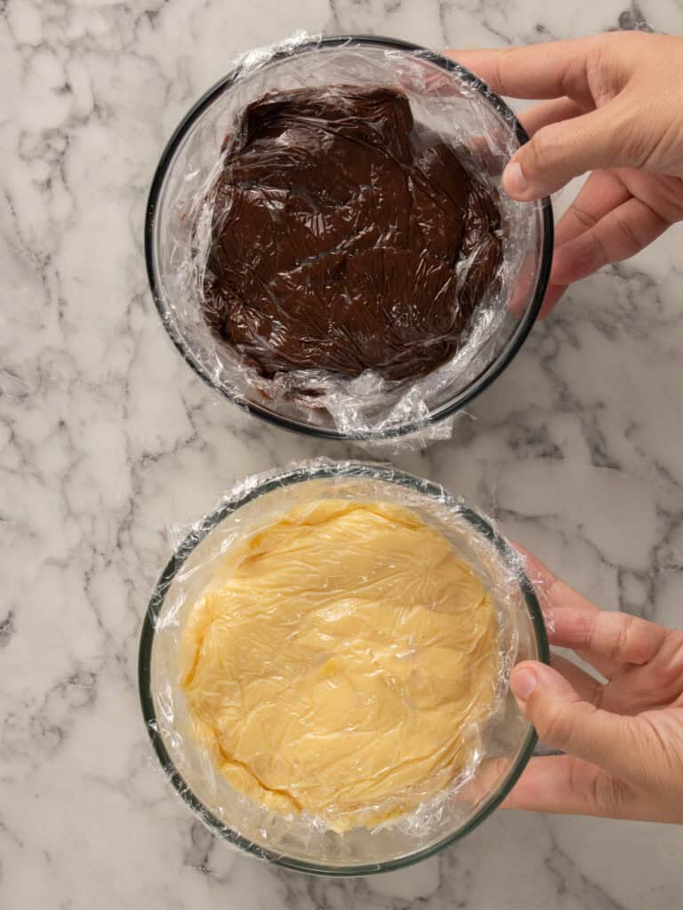 Two hands holding glass bowls on a marble surface; one bowl contains chocolate custard, the other contains vanilla custard, both covered with plastic wrap.