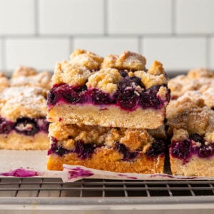 Two blueberry crumb bars stacked on top of each other, showing layers of golden crust, juicy blueberry filling, and crumbly topping, with more bars and a cooling rack in the background.