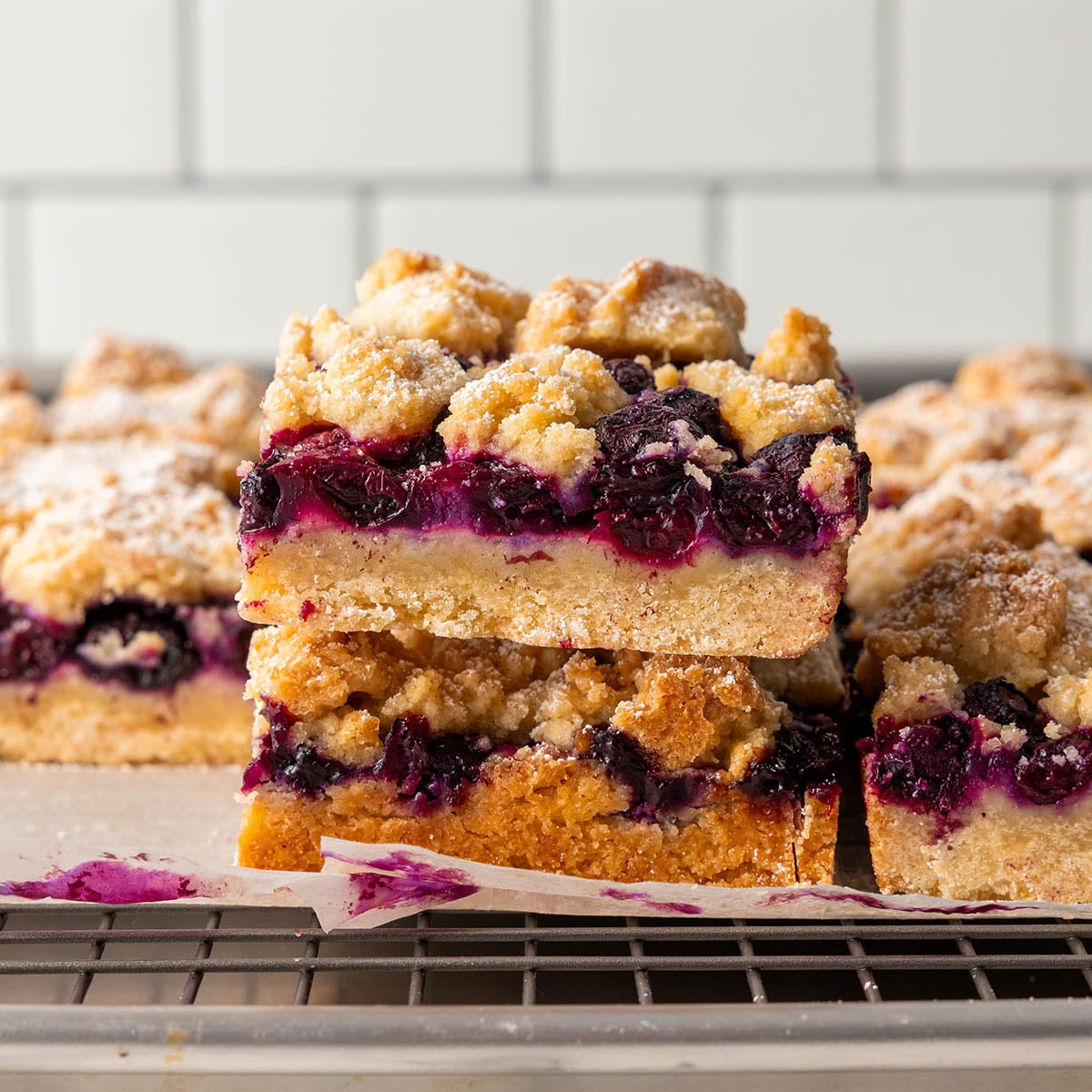 Two blueberry crumb bars stacked on top of each other, showing layers of golden crust, juicy blueberry filling, and crumbly topping, with more bars and a cooling rack in the background.