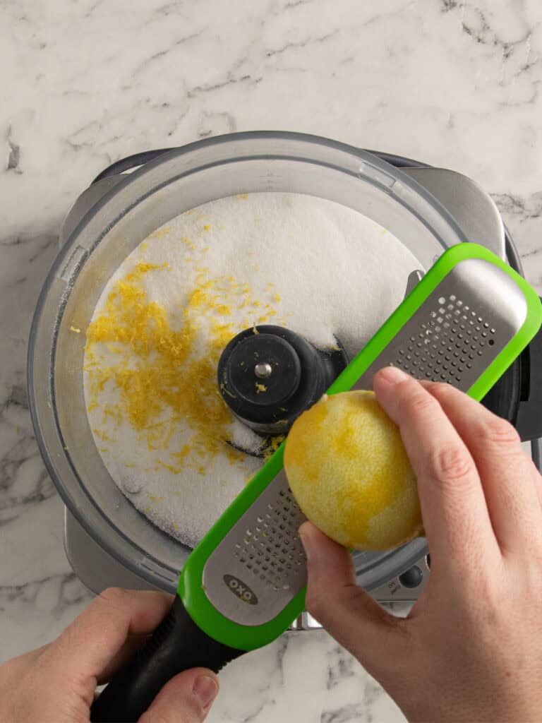 A hand uses a green zester to grate lemon zest into a food processor filled with sugar, set on a marble countertop.