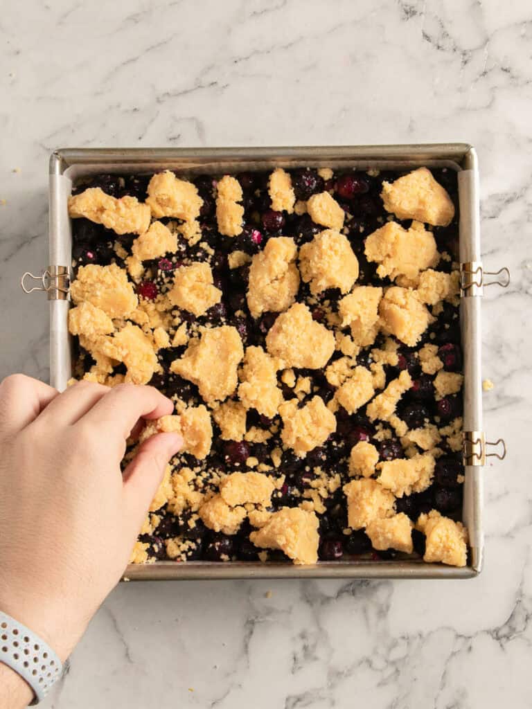 A hand sprinkles crumbly dough topping over a square baking pan filled with blueberries on a marble countertop.