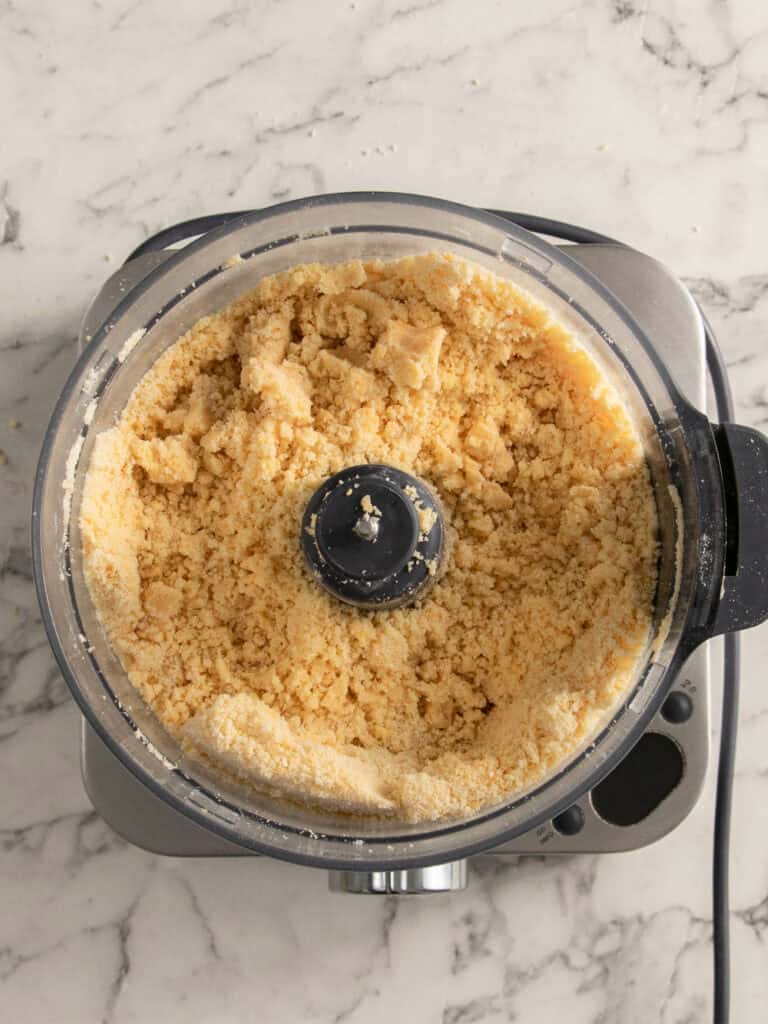 A top-down view of a food processor on a marble countertop, filled with coarse, crumbly dough mixture.