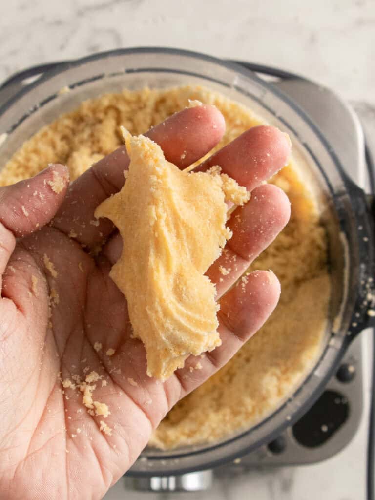 A hand holds a small amount of crumbly, yellow dough over a food processor bowl filled with more dough, on a light countertop background.
