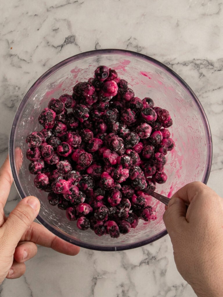 A person stirs blueberries coated in flour and sugar in a clear glass mixing bowl on a light marble countertop.