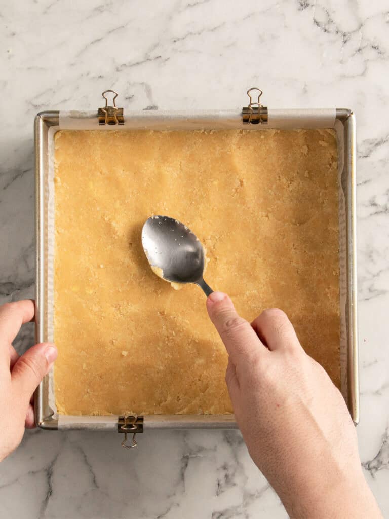 A person uses a spoon to press a crumbly dough evenly into a square baking pan lined with parchment paper on a marble countertop.
