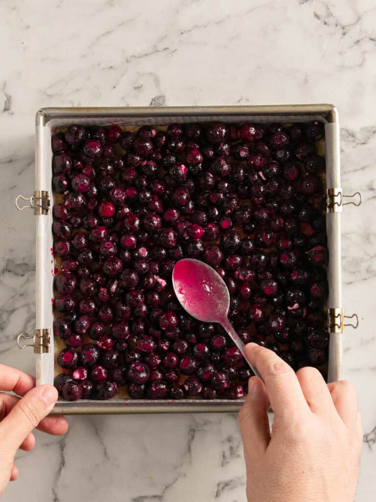 A person spreads a layer of fresh blueberries in a square metal baking pan with a spoon, on a marble countertop.
