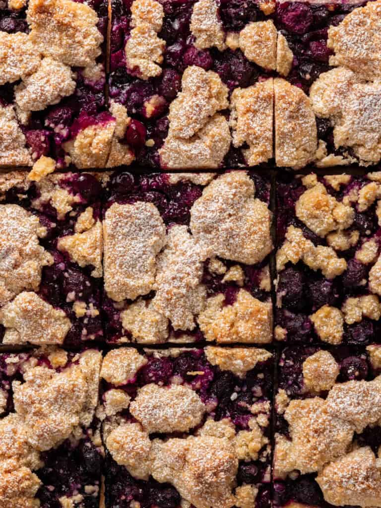 Close-up of baked berry crumble bars cut into squares, with a crumbly golden topping and a dusting of powdered sugar over a dark berry filling.