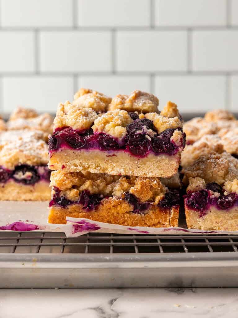 Two stacked blueberry crumb bars with a golden, crumbly topping and vibrant purple blueberry filling, resting on a wire cooling rack. More bars are in the background, with a white subway tile wall behind them.