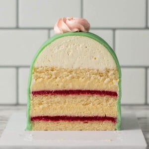 A square image of a cross-section of a Swedish princess cake with green icing, a dome-shaped top, and a pink meringue. The cake has layers of sponge, cream, and bright red jam, set against a white tiled background.