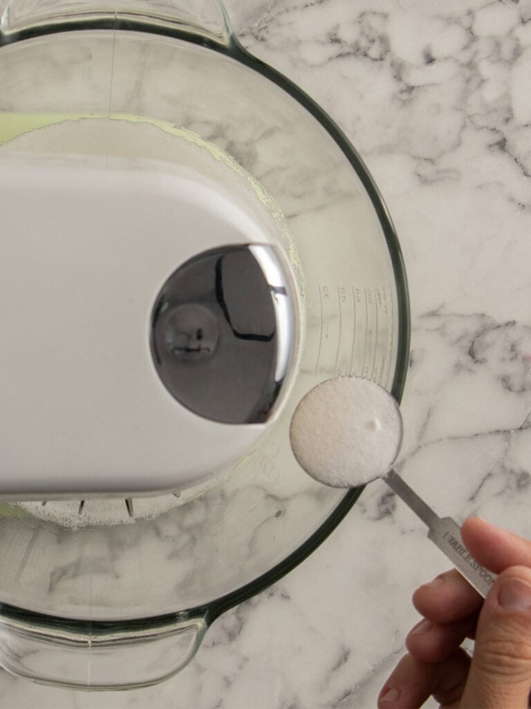 A hand holds a tablespoon of sugar above a glass mixing bowl with a stand mixer on a marble countertop, preparing to add the sugar to the bowl.