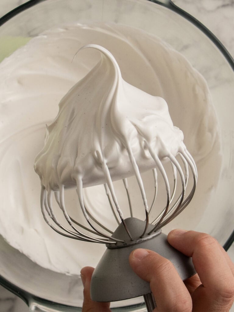 A hand holds a metal whisk attachment coated with thick, glossy white meringue, with more meringue visible in a glass mixing bowl below.