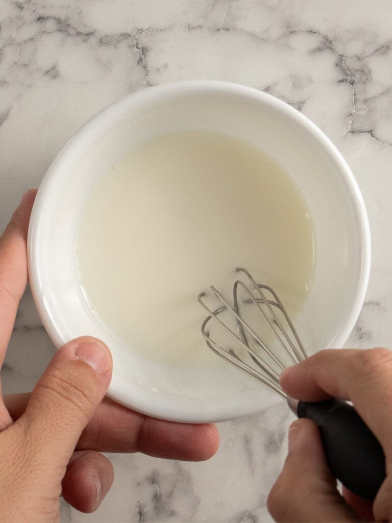 A person whisking a corn flour and water liquid in a small white bowl on a marble countertop.