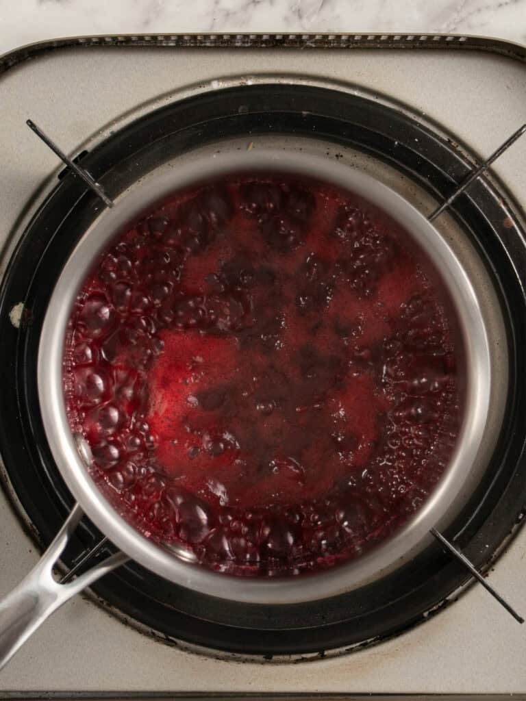 A saucepan filled with bubbling red raspberry jam sits on a stovetop burner.