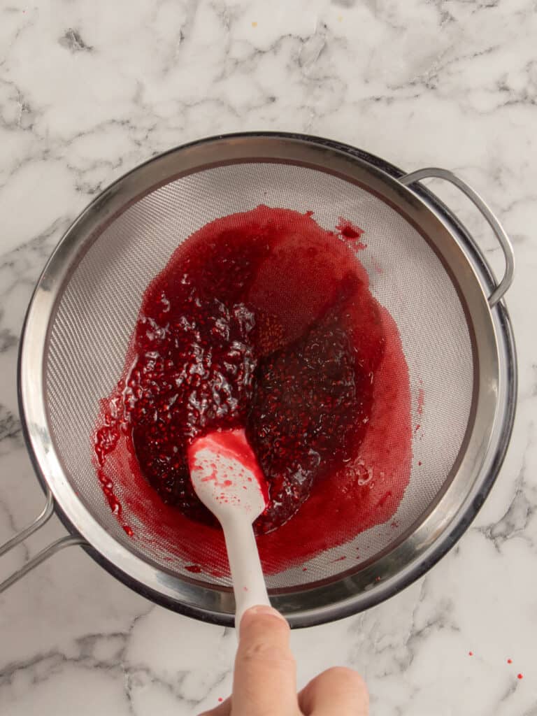 A hand uses a white spatula to press red raspberry sauce through a fine mesh strainer over a metal bowl, removing seeds. The background is a white marble countertop.