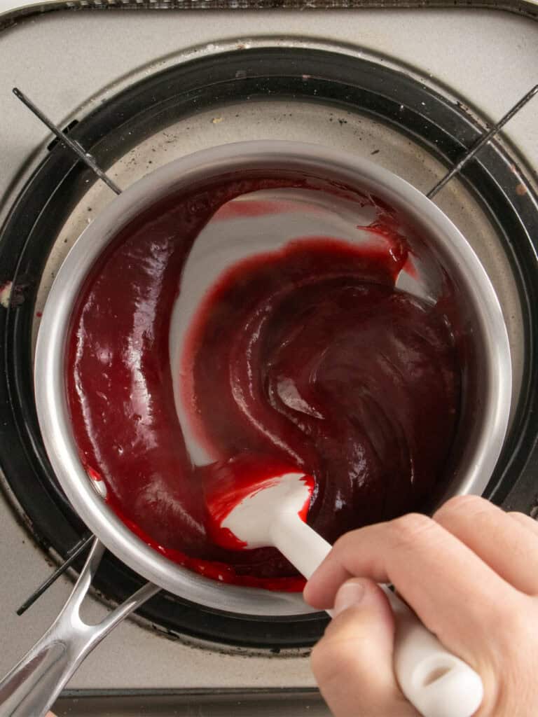 A hand stirs a thick, glossy red raspberry sauce in a saucepan on a stovetop using a white spatula.