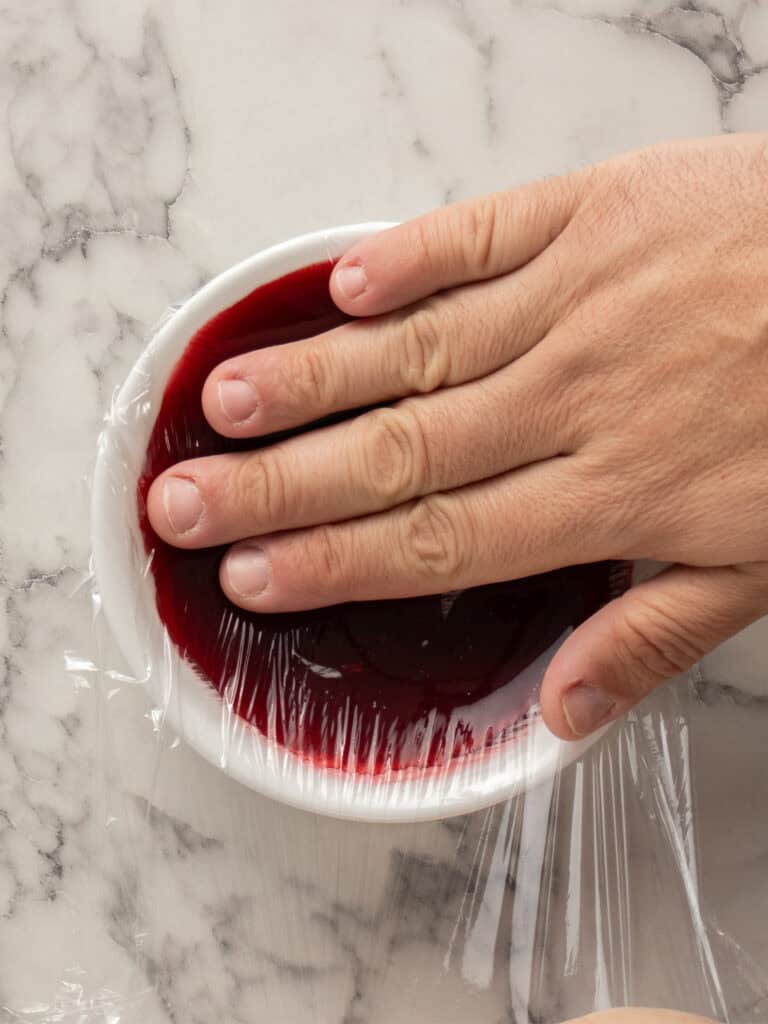 A hand pressing plastic wrap tightly over a white bowl filled with raspberry sauce, on a marble surface.