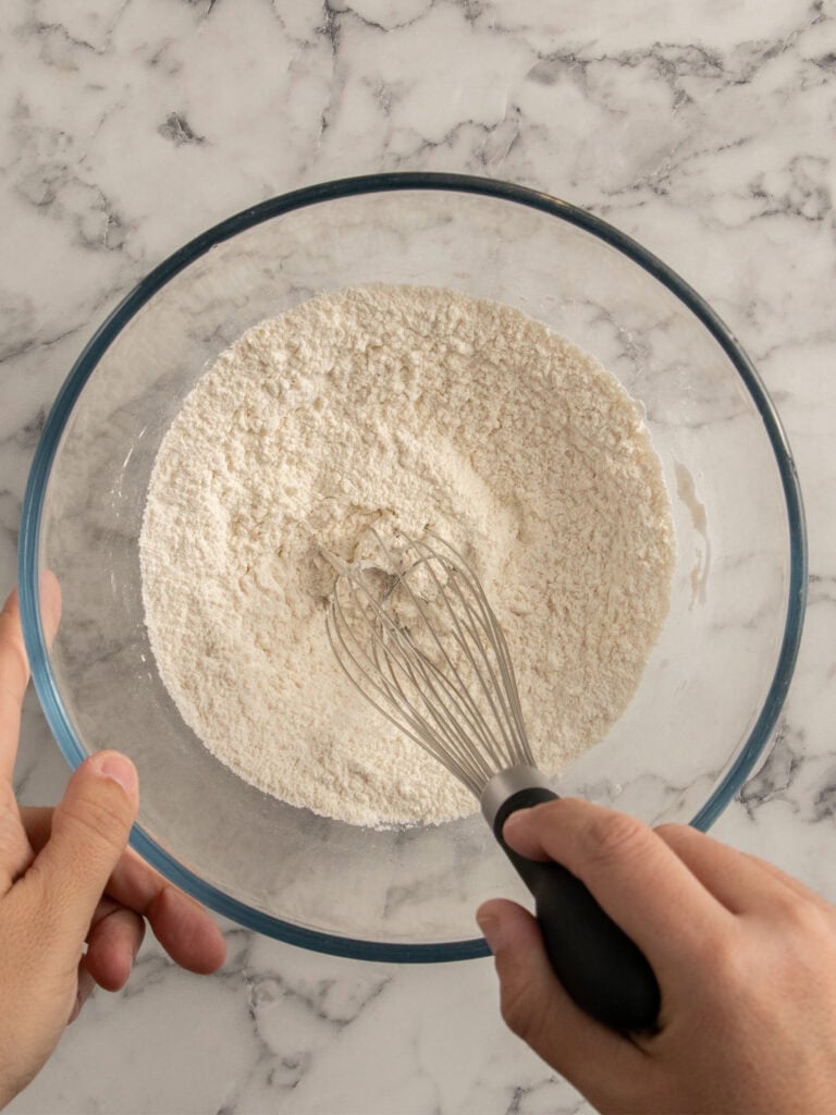 A person holding a glass bowl of flour and using a whisk to mix the dry ingredients, on a marble countertop.