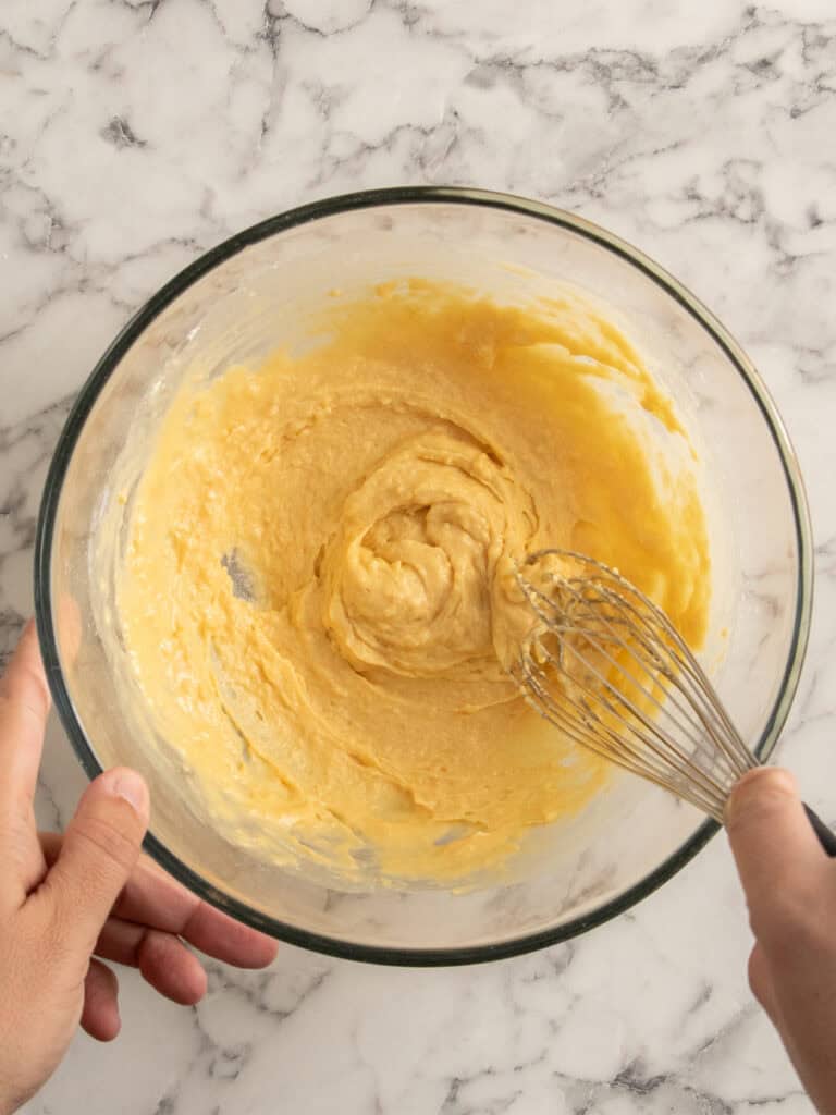 A persons hands are holding a glass bowl with yellow cake batter being mixed with a metal whisk on a white marble surface.